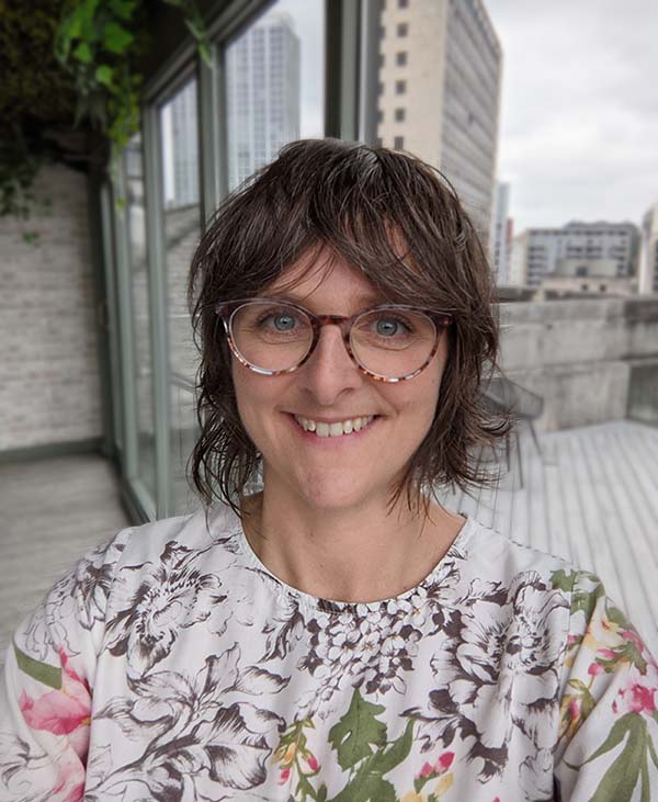 Photo of a coach with short brown hair and glasses. She's smiling at the camera with a city scape behind her.